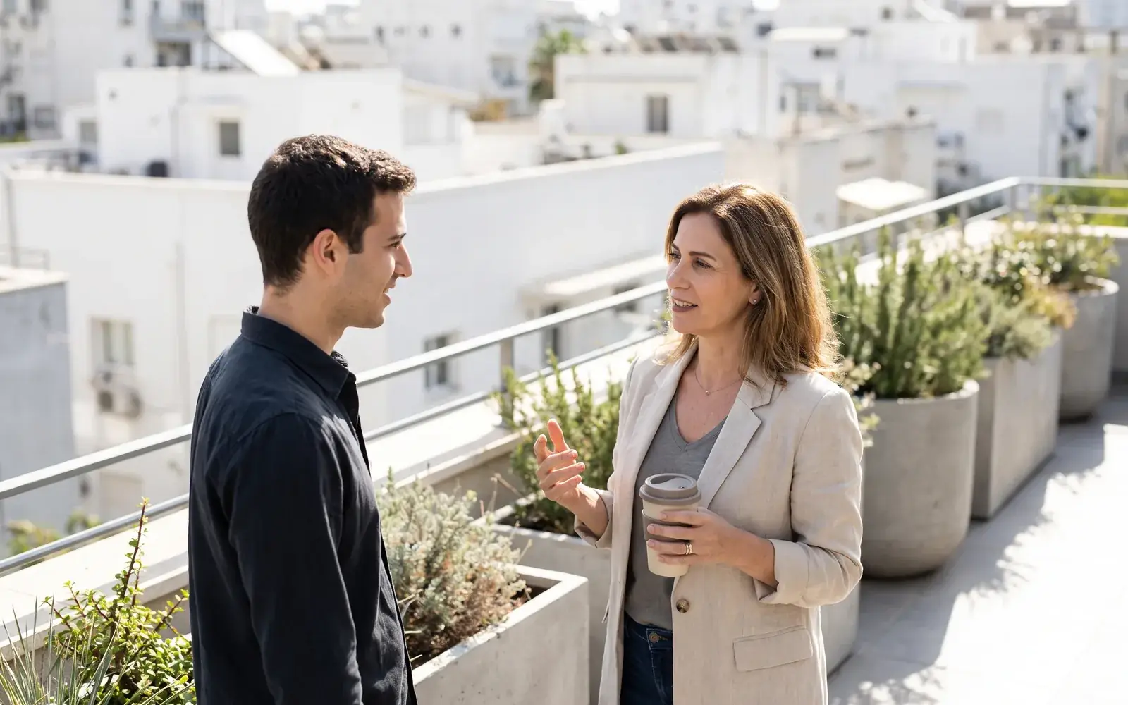 Two tech executives talk with coffee on a sunlit rooftop terrace of an office building, white city blocks behind